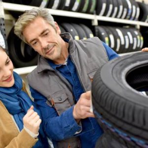 A man and a woman look at a tire in a shop. The man is touching the tire and pointing at the tag on it.