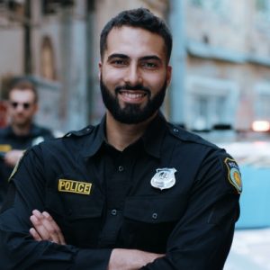 A male police officer wearing a blue uniform is crossing his arms and smiling in front of a police car.