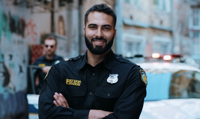 A male police officer wearing a blue uniform is crossing his arms and smiling in front of a police car.