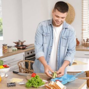A close-up of a young man in the kitchen cutting vegetables for his lunch box. He's wearing a denim shacket and smiling.