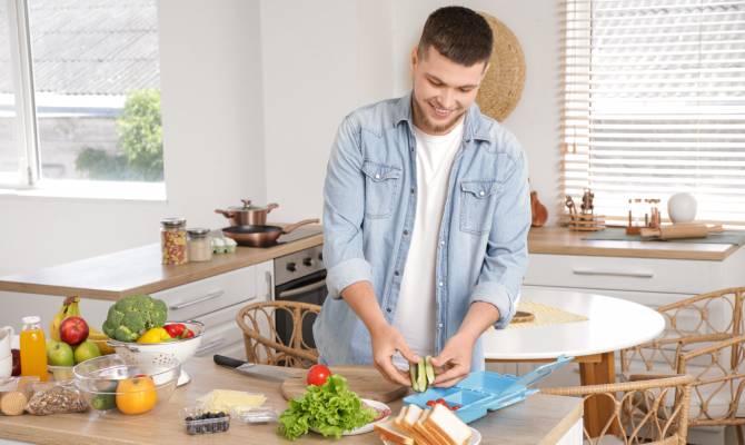 A close-up of a young man in the kitchen cutting vegetables for his lunch box. He's wearing a denim shacket and smiling.