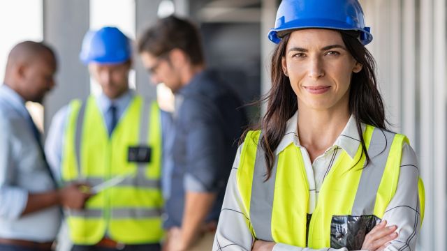 A female contractor standing in the foreground with her arms crossed and three males blurred out in the background.