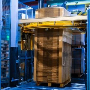 A large, blue manufacturing machine is wrapping a stack of flattened cardboard boxes with plastic wrap.