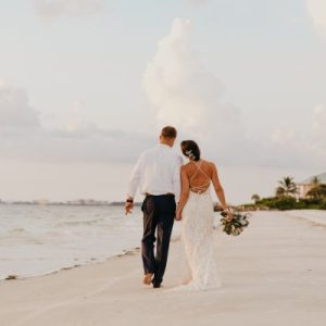 A man wearing a white shirt and black pants holds hands with a woman wearing a form-fitting wedding dress on the beach.