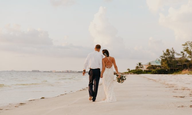 A man wearing a white shirt and black pants holds hands with a woman wearing a form-fitting wedding dress on the beach.