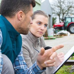 A woman holds a clipboard as she and a man kneel near a piece of heavy equipment. A man and a tractor are in the background.