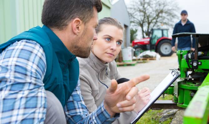 A woman holds a clipboard as she and a man kneel near a piece of heavy equipment. A man and a tractor are in the background.