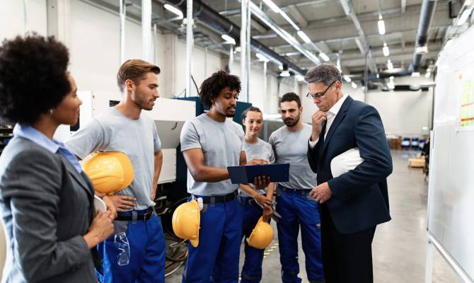 A man and woman in business suits speak with four people wearing blue pants and gray shirts in a factory.