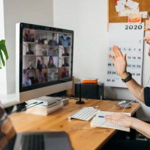 A remote working employee sitting in front of a laptop and monitor. He's wearing headphones and waving at the screen.
