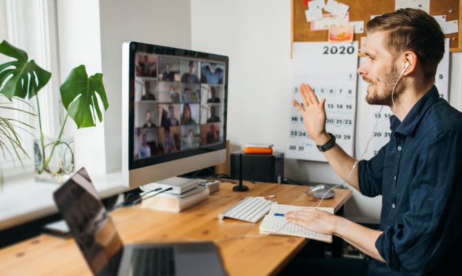 A remote working employee sitting in front of a laptop and monitor. He's wearing headphones and waving at the screen.