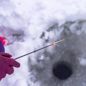 The hand of a gloved angler holding a small fishing rod over a hole in the ice. Their pants and gloves are purple.