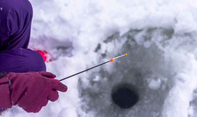 The hand of a gloved angler holding a small fishing rod over a hole in the ice. Their pants and gloves are purple.