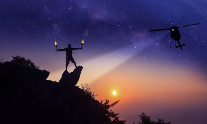 A helicopter shining a light toward the ground in the evening. A person on the ground is holding up a fire signal.