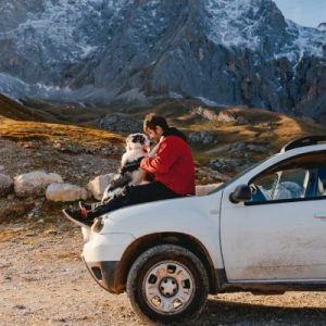 A man and his dog sitting on the hood of his off-roading vehicle with a mountainous landscape in the background.