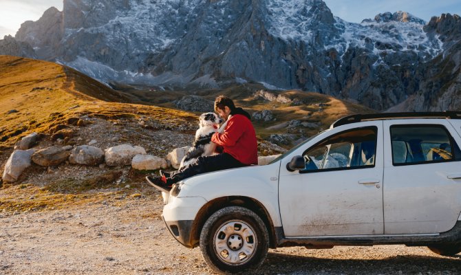 A man and his dog sitting on the hood of his off-roading vehicle with a mountainous landscape in the background.
