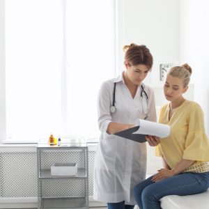 A female doctor wearing a white coat is holding a clipboard with paperwork and reviewing it with a female patient.
