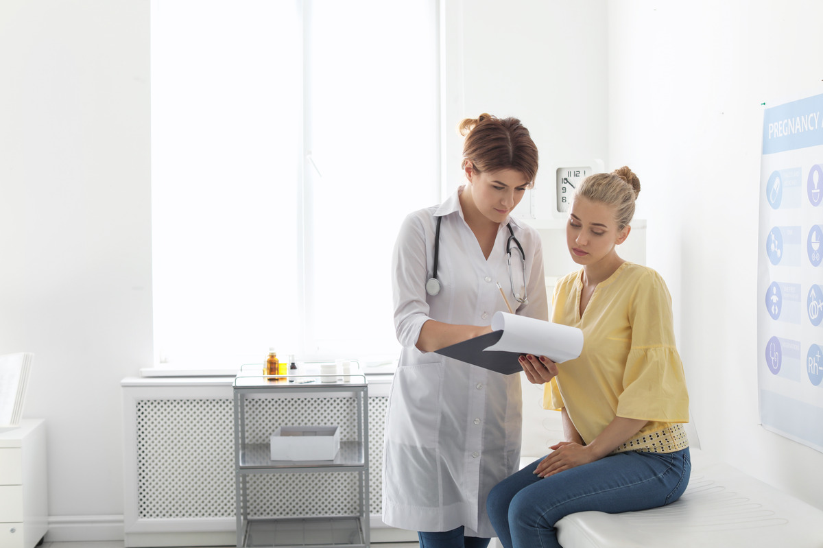 A female doctor wearing a white coat is holding a clipboard with paperwork and reviewing it with a female patient.