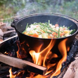 A black pot sitting over a campfire. There are vegetables inside of the pot and there's grass in the background.