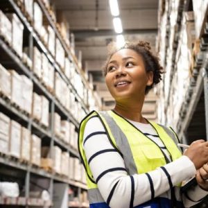 A woman in a striped shirt and bright yellow safety vest stands in a warehouse aisle, writing on a notepad.