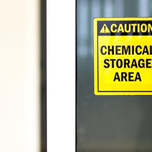 A yellow and black sign with a picture of a barrel that says "Caution: Chemical Storage Area" is seen stuck to glass.