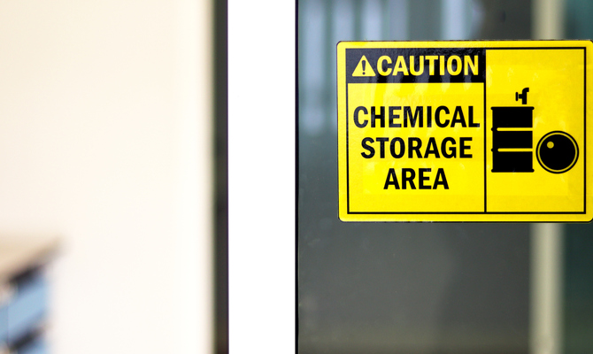 A yellow and black sign with a picture of a barrel that says "Caution: Chemical Storage Area" is seen stuck to glass.