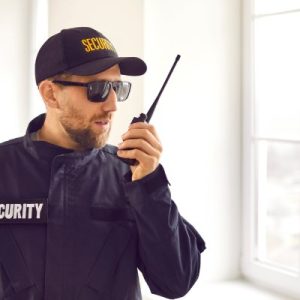 A man in a security uniform and sunglasses speaking into a two-way radio in a white room with a window.