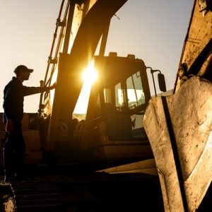 A person wearing a baseball cap has climbed onto a large excavator. The sun shines through the arm of the equipment.