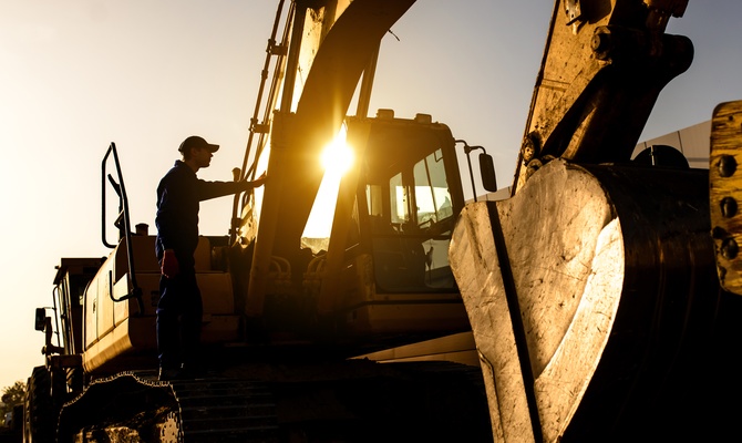 A person wearing a baseball cap has climbed onto a large excavator. The sun shines through the arm of the equipment.
