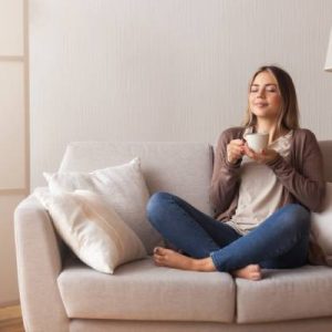 A young woman holds a mug, sits cross-legged on a loveseat in her living room, and smiles peacefully.