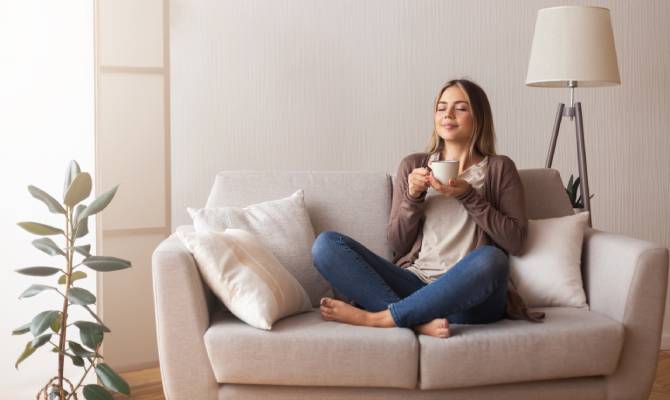 A young woman holds a mug, sits cross-legged on a loveseat in her living room, and smiles peacefully.