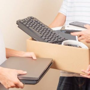 One person is holding a box with electronics. Another person places phones in the box, and a third person holds laptops.