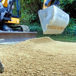 A compact excavator dumps a load of fresh gravel on a driveway. A worker nearby holds a tool to level the surface.