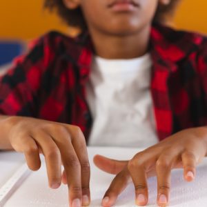 A close-up of a young boy in a plaid shirt sitting at a desk while using both hands to read a braille book.