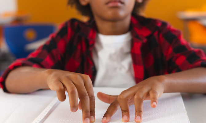 A close-up of a young boy in a plaid shirt sitting at a desk while using both hands to read a braille book.