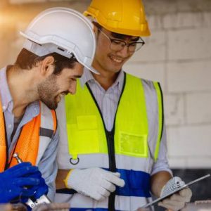 Two men wearing hard hats, vests, and safety gloves look at something on a tablet while working in a facility.
