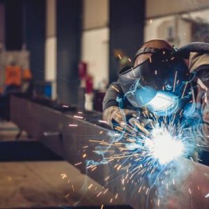 An industrial welder wearing a mask and gloves uses a torch on a long steel beam. Blue and gold sparks fly off the metal.