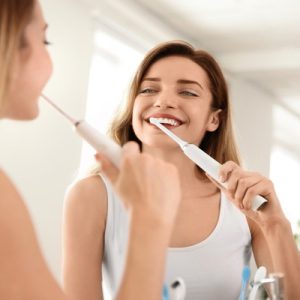 A woman wearing a white tank top is looking at herself in a mirror while brushing her teeth with an electric toothbrush.