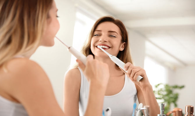 A woman wearing a white tank top is looking at herself in a mirror while brushing her teeth with an electric toothbrush.