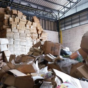 A messy, chaotic warehouse with piles of discarded boxes on the ground and towering, unstable stacks of boxes against the wall.