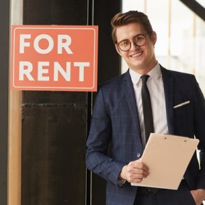 A man wearing a suit stands next to a sign that says, "For Rent." He's smiling while holding a clipboard.