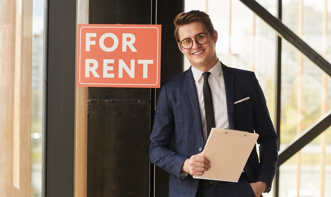 A man wearing a suit stands next to a sign that says, "For Rent." He's smiling while holding a clipboard.