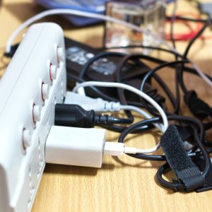 Cables from multiple devices plugged into a power strip on top of a table. The tangled cables are behind a laptop.
