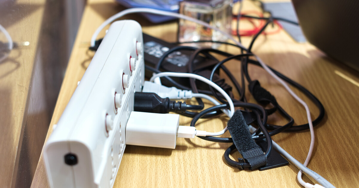 Cables from multiple devices plugged into a power strip on top of a table. The tangled cables are behind a laptop.