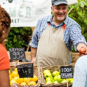 A smiling farmer passing an apple to a patron of his farm stand. Another patron looks at the display.