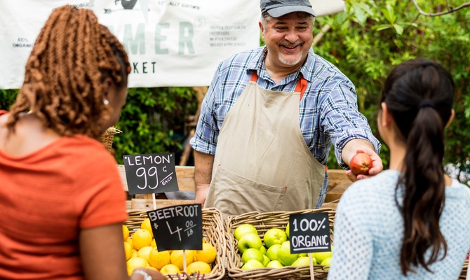 A smiling farmer passing an apple to a patron of his farm stand. Another patron looks at the display.