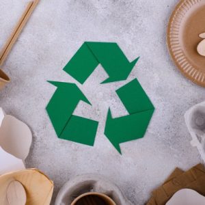 Reusable food packaging materials like egg cartons, cardboard plates, cups, and wooden spoons on a grey surface with a green recycling sign in the center.