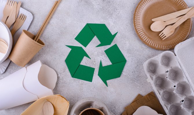 Reusable food packaging materials like egg cartons, cardboard plates, cups, and wooden spoons on a grey surface with a green recycling sign in the center.