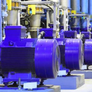 Various large blue electric generators inside of a factory aligned in a single row with various connections and piping.