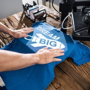 A person's arms holding a blue T-shirt in place on a DTF heat press to transfer a rocket design that reads "Dream Big."