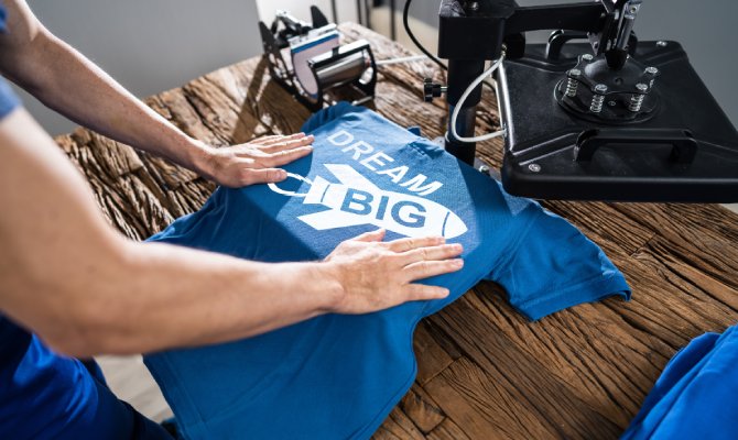 A person's arms holding a blue T-shirt in place on a DTF heat press to transfer a rocket design that reads "Dream Big."